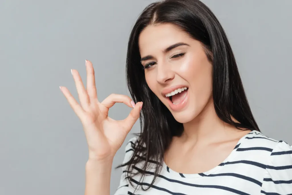 Playful young woman standing over grey background while make Ok gesture. Look at camera.